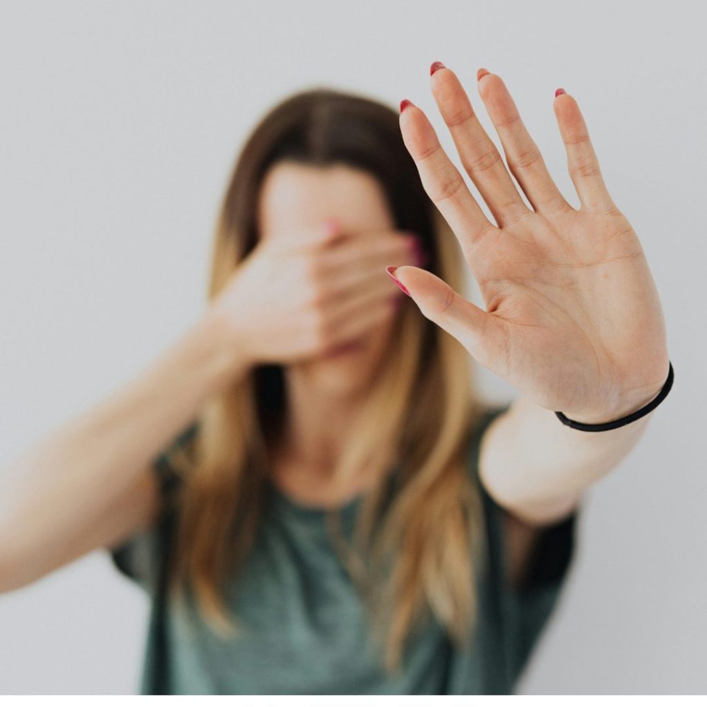 A woman with long, light brown hair is holding up her hand with her palm facing the camera, as if signaling "stop." She is slightly blurred in the background, with her other hand covering her face. The background is plain and light-colored.