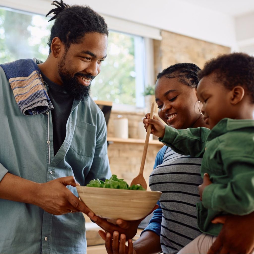 A man with dreadlocks and a beard smiling while holding a bowl of salad, engaging happily with a woman and a young child in a bright, cozy kitchen. The woman is smiling and holding a wooden spoon, while the child is reaching towards the bowl with curiosity.
