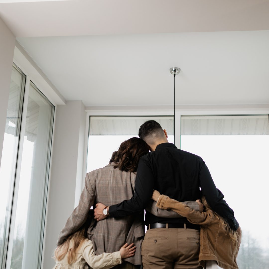 A family of four standing close together indoors, with their arms around each other, facing a large window. Their backs are to the camera, and they appear to be sharing a tender moment.