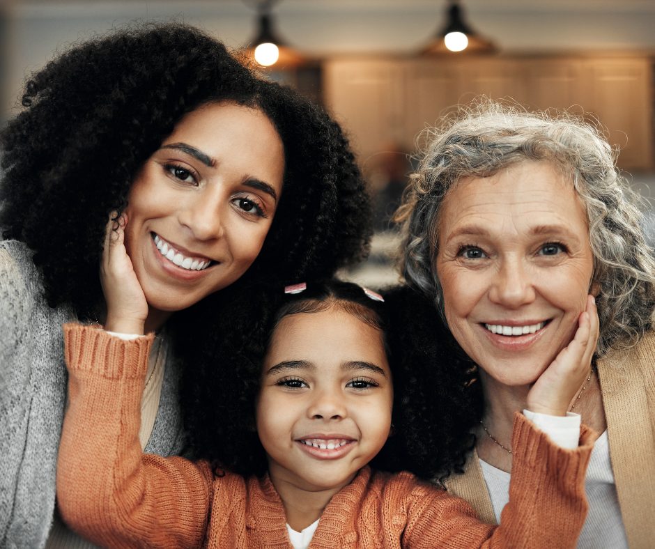 A happy multigenerational family of three, including a young girl, a woman with dark curly hair, and an older woman with gray curly hair, all smiling and resting their heads on their hands. They are close together indoors, with a warm, cozy background.