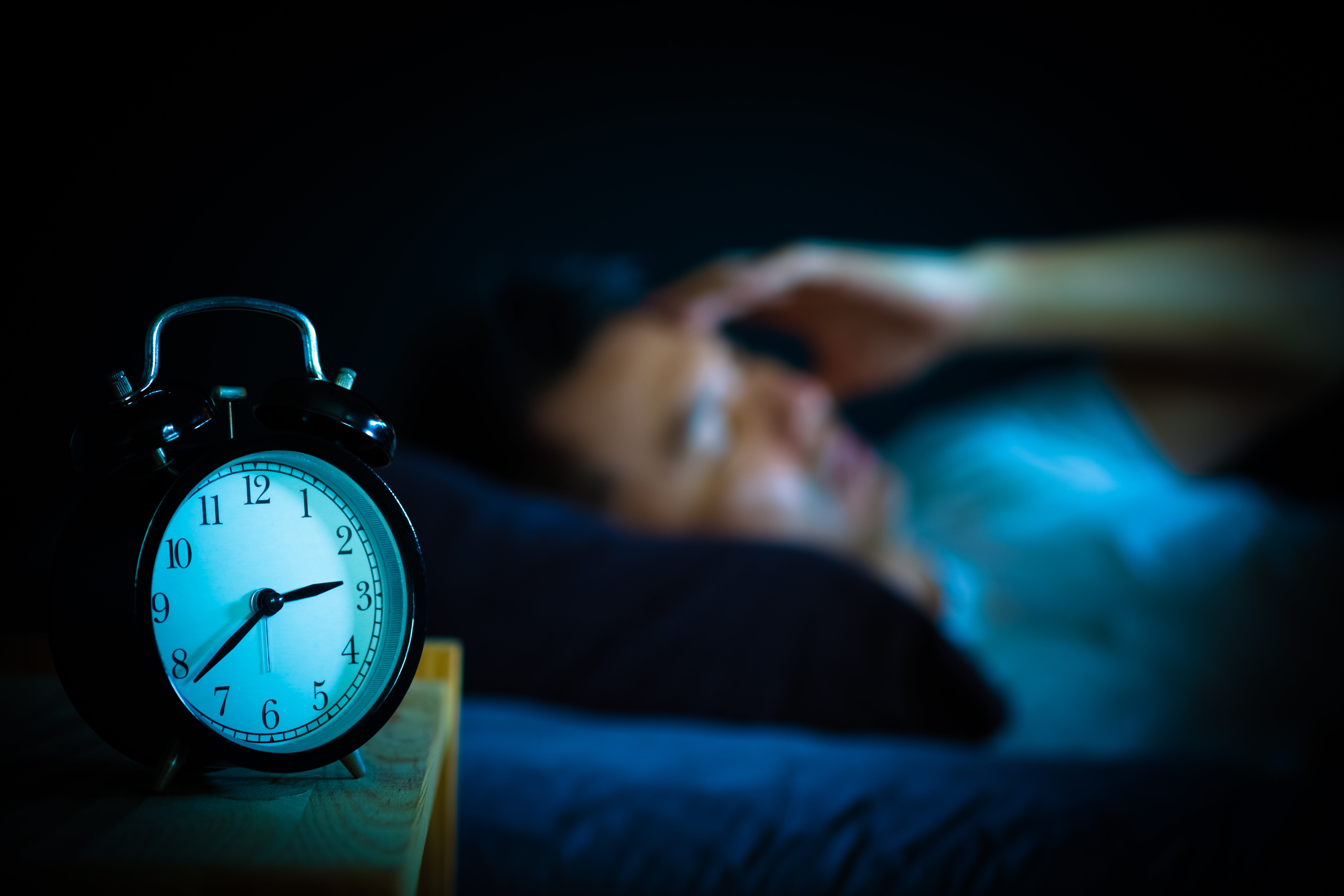 Blurred image of a man lying awake in bed at night, holding his head in frustration, with a clear view of an alarm clock in the foreground showing the late hour.