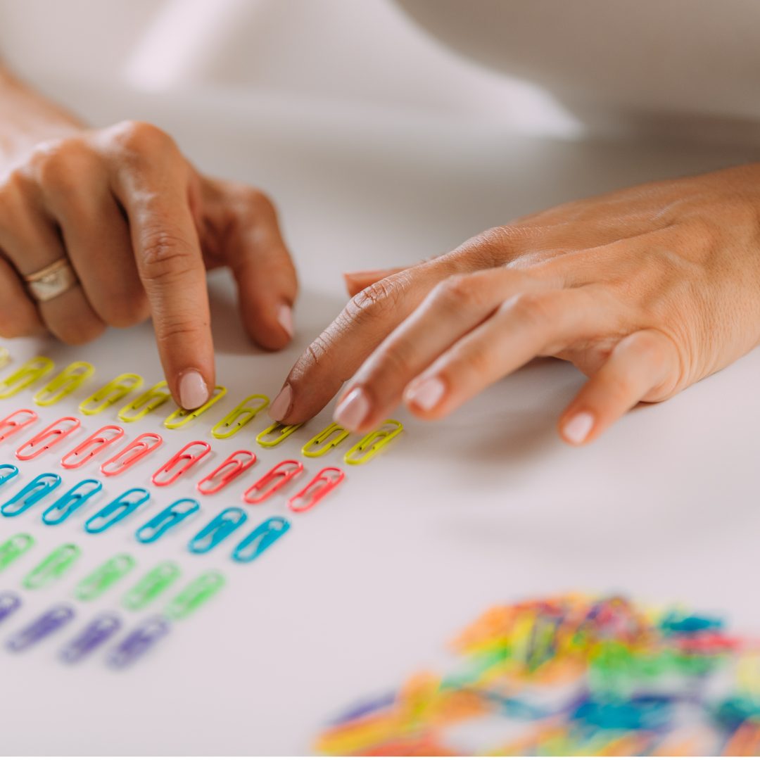 A close-up of a person's hands arranging colorful paper clips on a white surface. The person is wearing a ring on their left ring finger, and their nails are neatly manicured. There are additional paper clips in various colors scattered at the bottom right corner of the image.