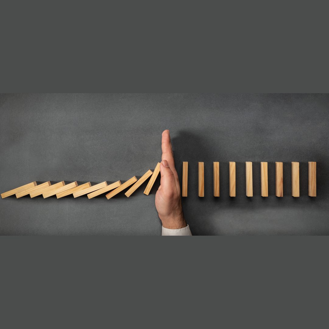 A hand stopping a series of wooden dominoes from falling, with the first set of dominoes knocked over and the second set standing upright, symbolizing intervention to prevent a chain reaction or collapse.