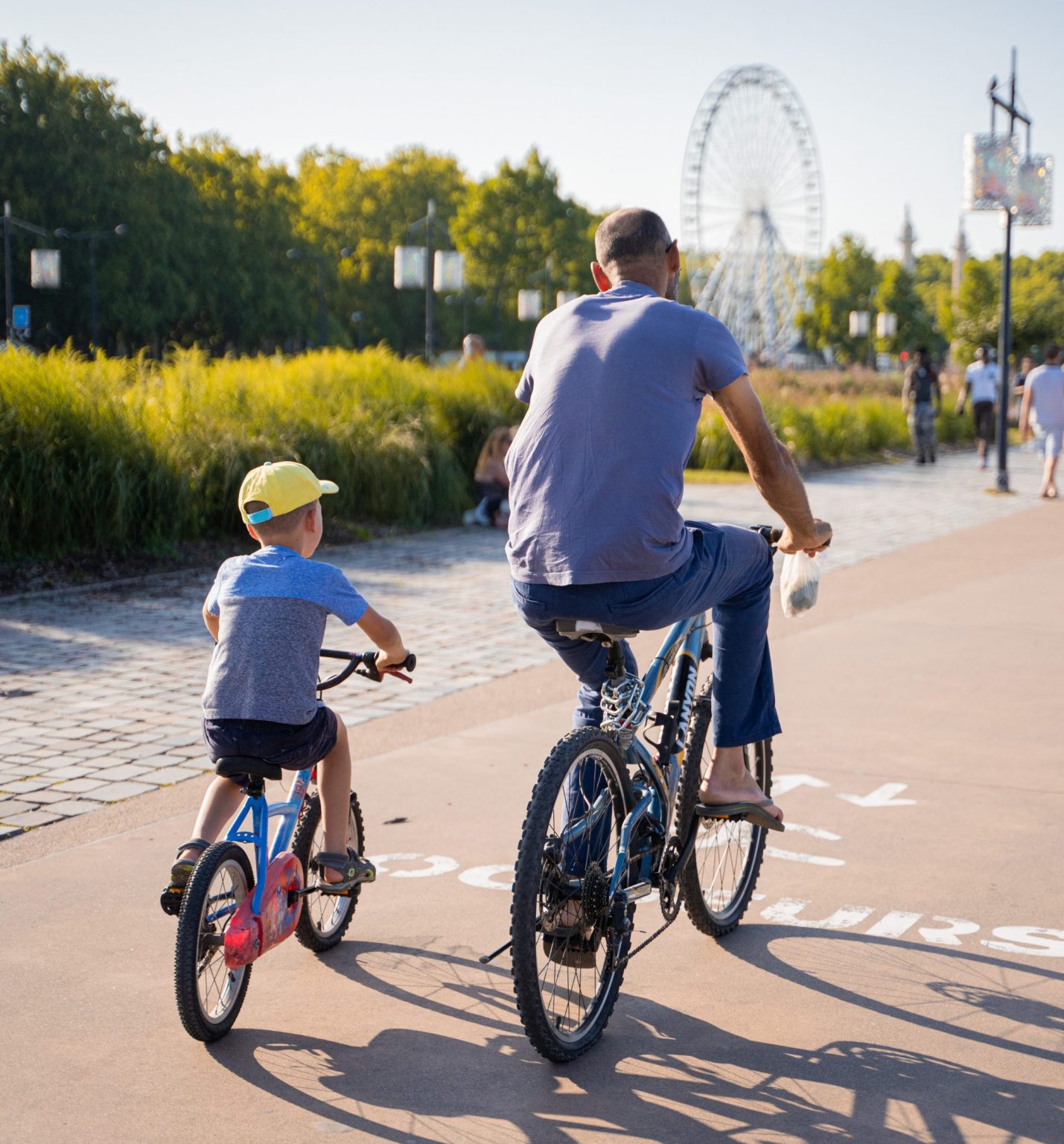 A man and a young boy are riding bikes together outdoors on a sunny day. The man is wearing a light blue t-shirt and dark blue pants, while the boy is wearing a gray t-shirt, dark shorts, and a yellow cap. They are riding on a paved path in a park, with greenery and trees in the background, along with a large Ferris wheel and other amusement park attractions visible in the distance.