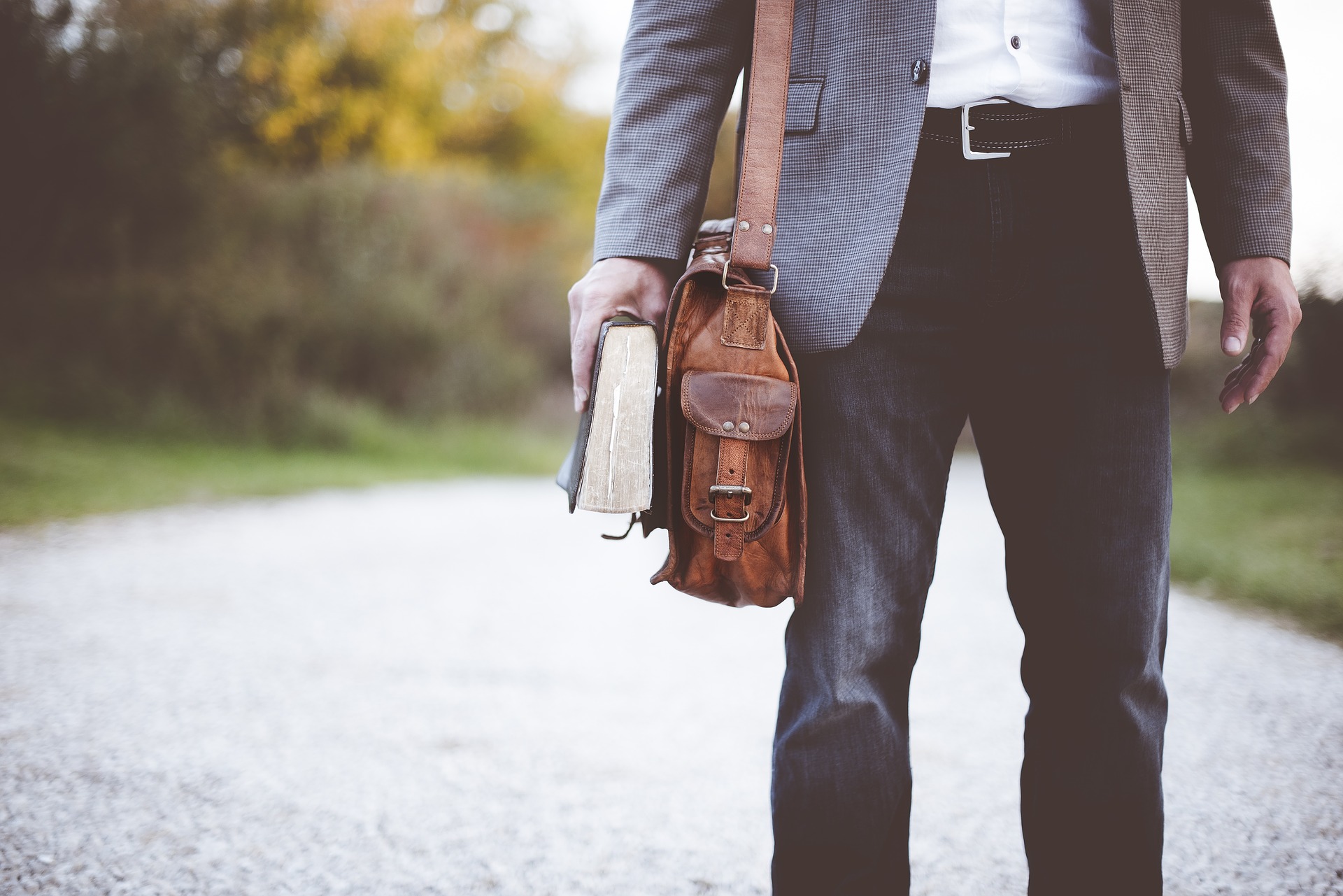 Man standing on a gravel path holding a worn book in one hand and a brown leather shoulder bag in the other, dressed in jeans and a blazer.