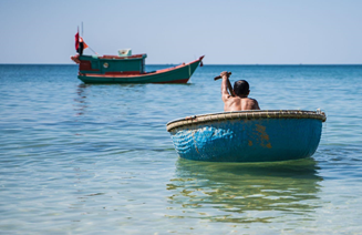Man paddling a small round blue boat in shallow, clear ocean water, heading toward a larger fishing boat floating in the distance.