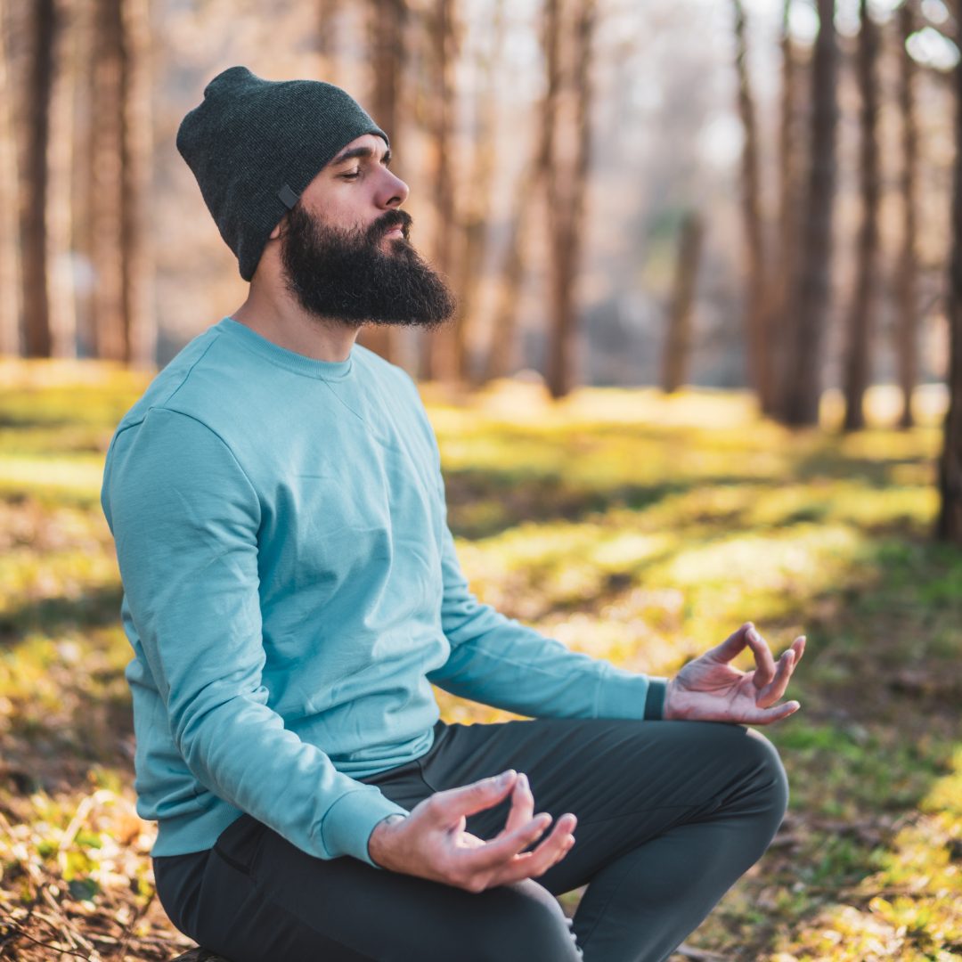 Man wearing a beanie and a long shirt is sitting down outside meditating.