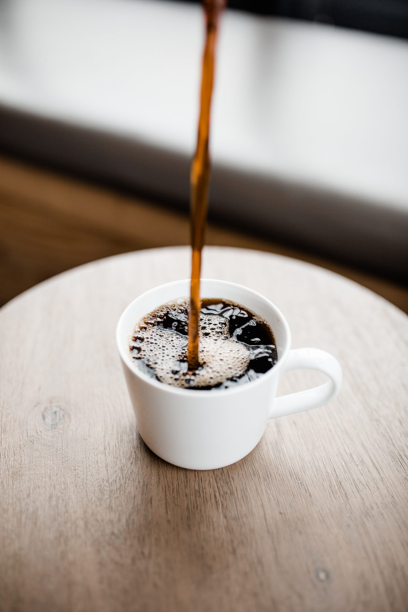A white ceramic mug placed on a wooden surface, with hot coffee being poured into it from above. The coffee creates bubbles and foam on the surface as it fills the mug. The background is blurred, emphasizing the focus on the mug and the stream of coffee.