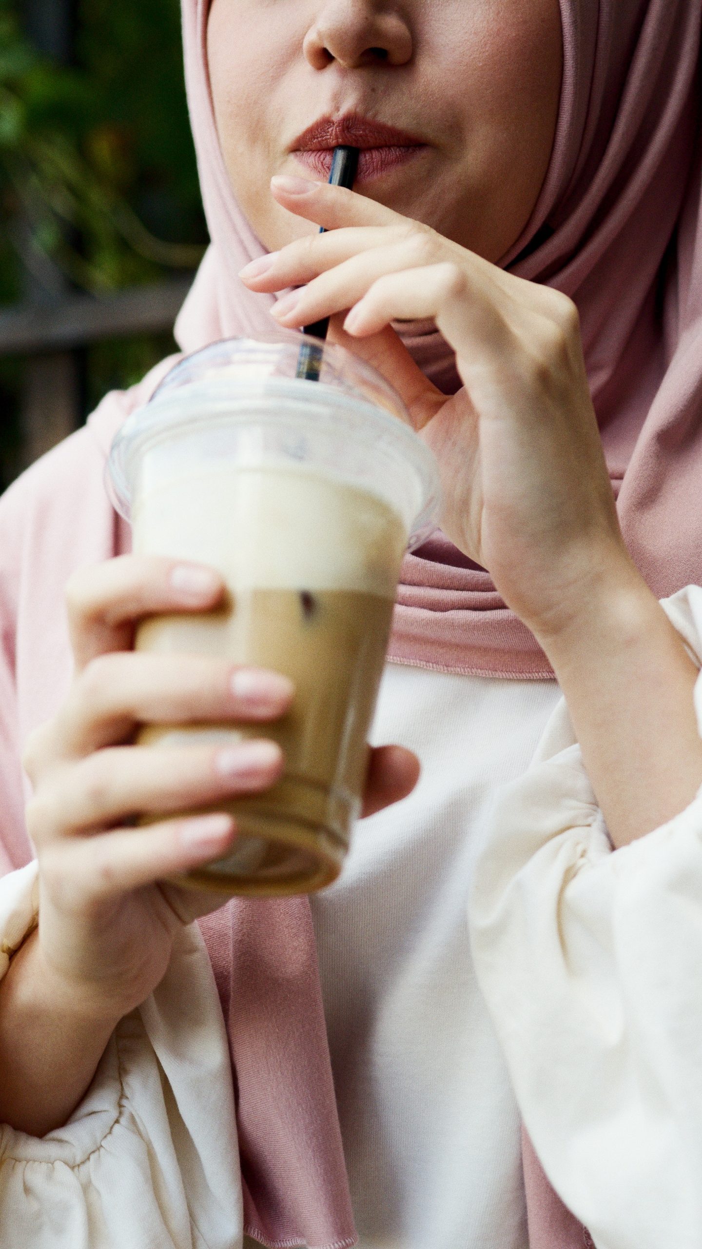 Close-up of a woman wearing a pink hijab, sipping a cold beverage through a black straw from a clear plastic cup with a lid, outdoors with blurred greenery in the background.