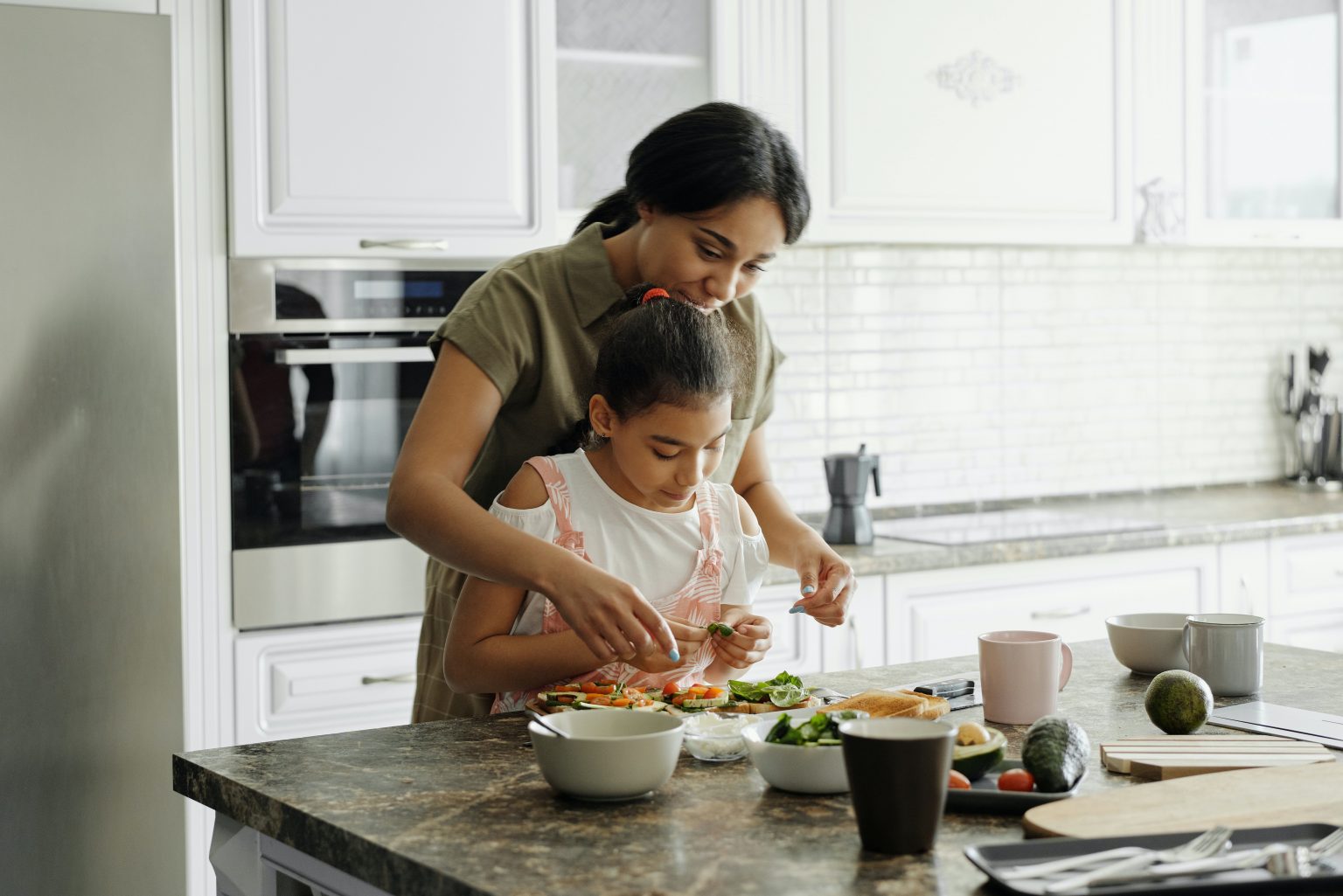 A woman and a young girl are preparing food together in a bright, modern kitchen. The woman is standing behind the girl, helping her as she arranges vegetables on a cutting board. The kitchen counter is filled with bowls, vegetables, and utensils, and the background features white cabinets, a built-in oven, and large windows letting in natural light.