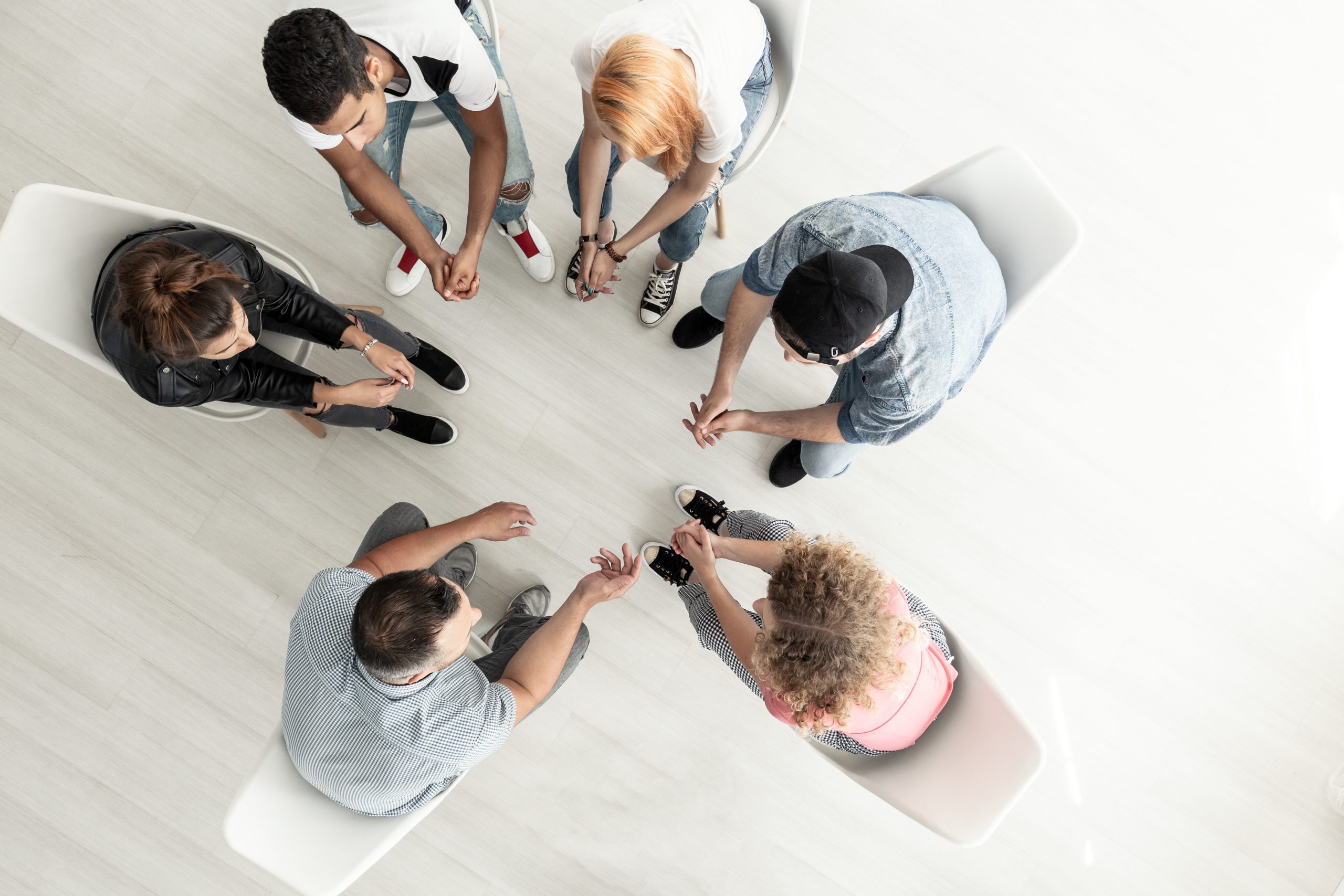 Six people sitting in a circle, holding hands and engaging in a group activity or prayer, viewed from above in a bright, minimalistic room with light-colored flooring and white chairs.