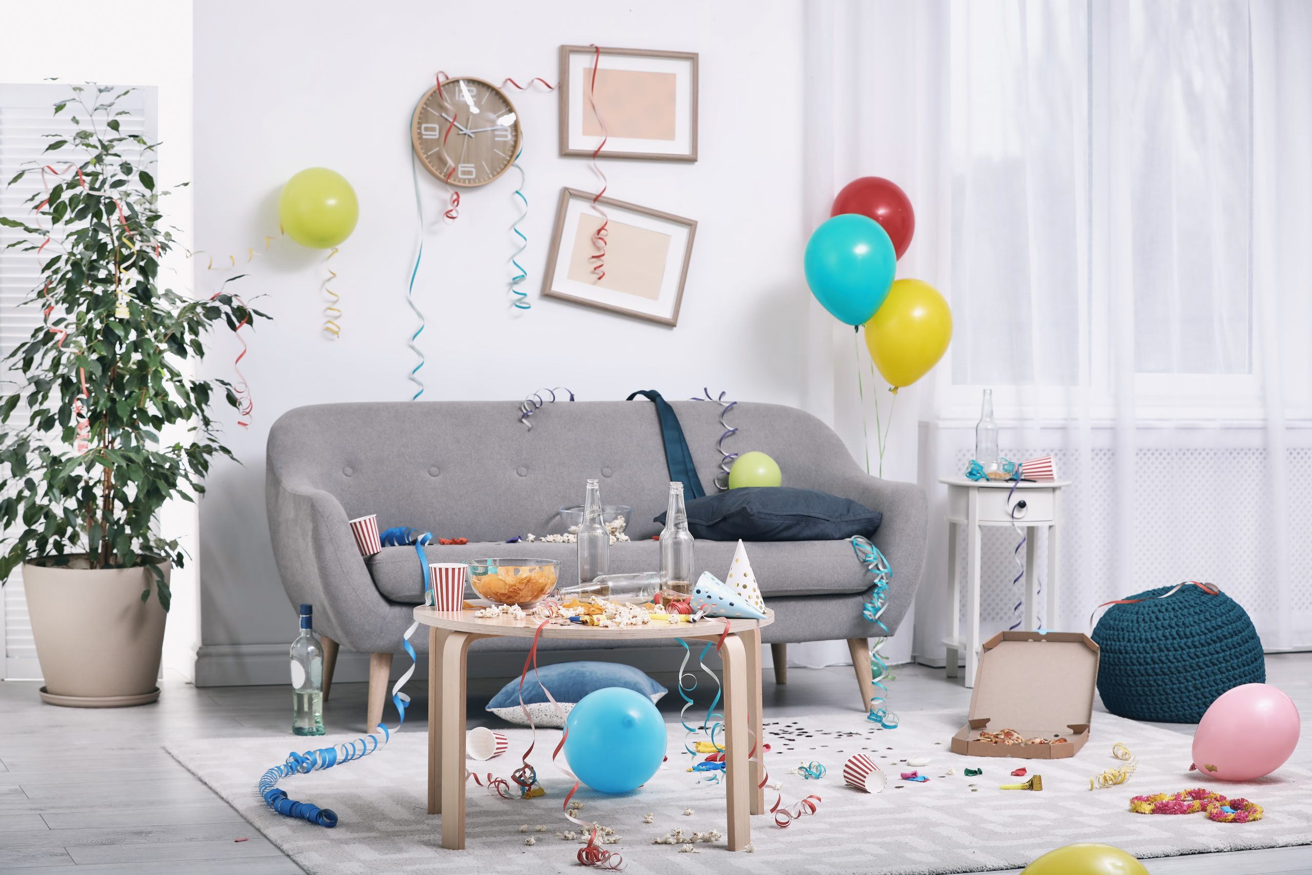 A messy living room decorated for a celebration with balloons, streamers, and party supplies. The gray sofa has a pillow and a balloon on it, while a small wooden table in front is cluttered with popcorn, snacks, and empty bottles. Balloons are scattered on the floor, and a cardboard pizza box is open with leftover pizza inside. Colorful streamers hang from the ceiling, and a potted plant is visible on the left side near a wall clock. The room has white walls, sheer curtains, and a cozy, festive atmosphere.