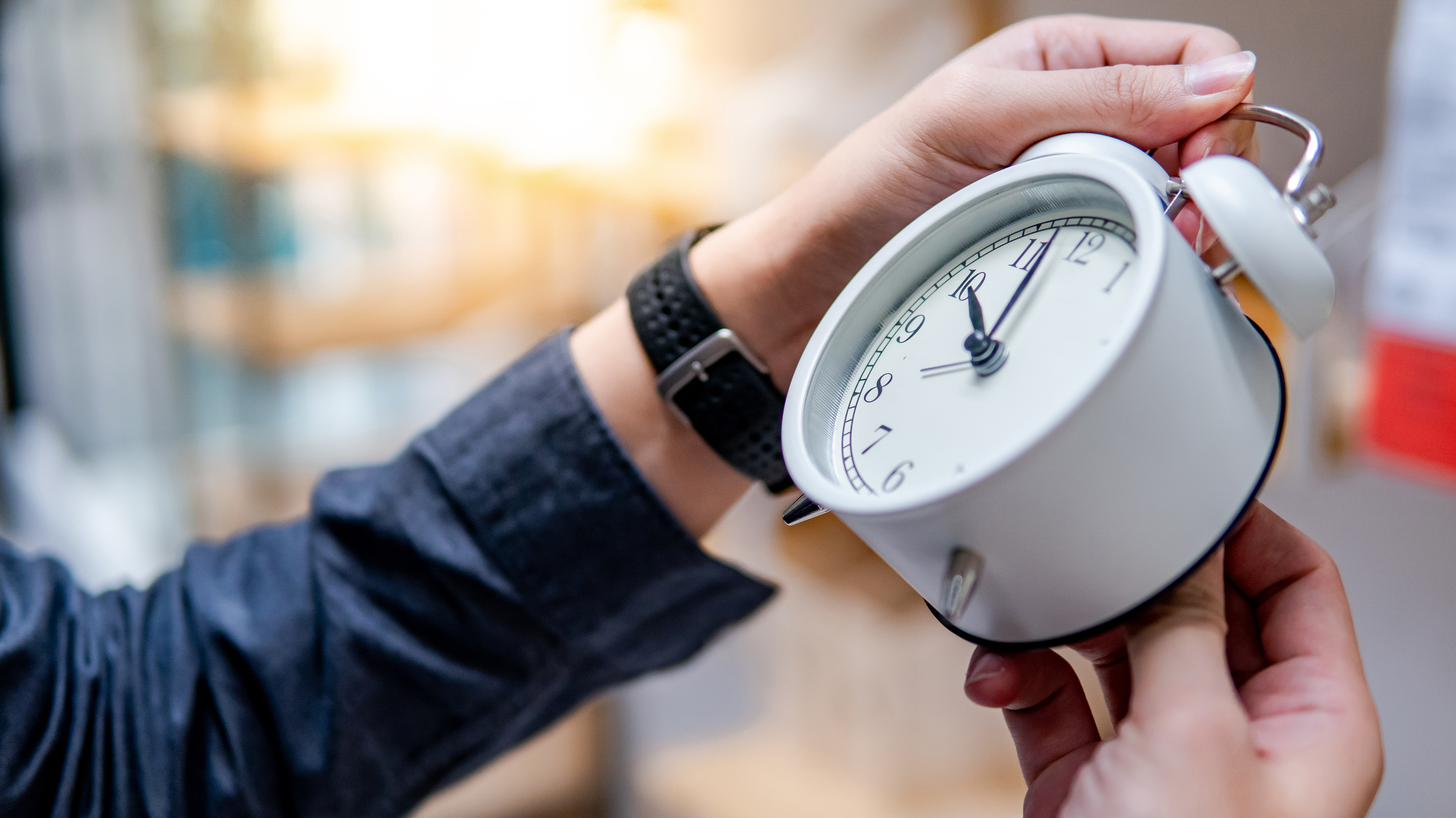 Close-up of hands holding and adjusting a white analog alarm clock, with a soft, warm background.