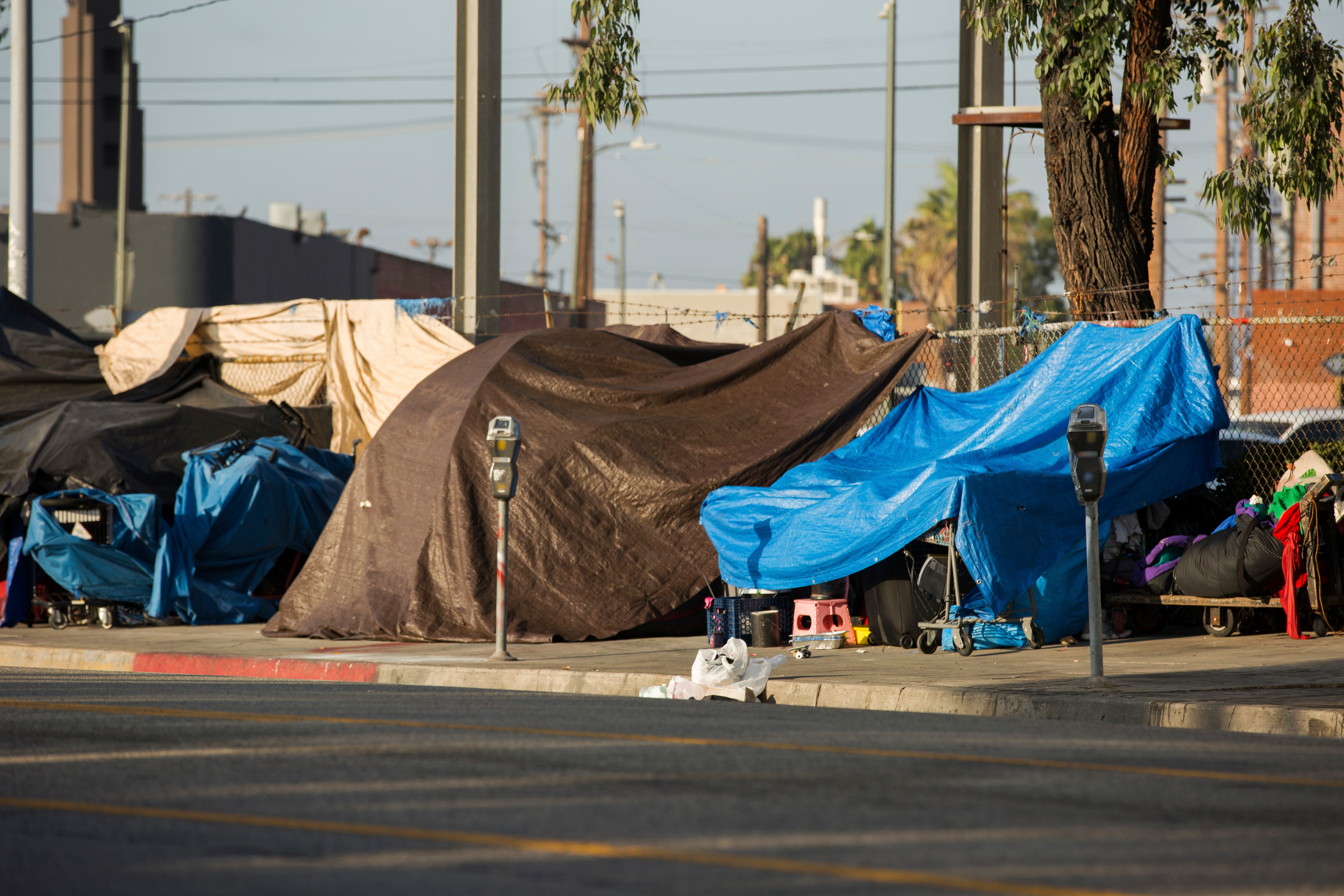 An outdoor scene showing a makeshift homeless encampment with tarps and blankets covering belongings and tents on the sidewalk, with a few scattered items and a chain-link fence in the background.