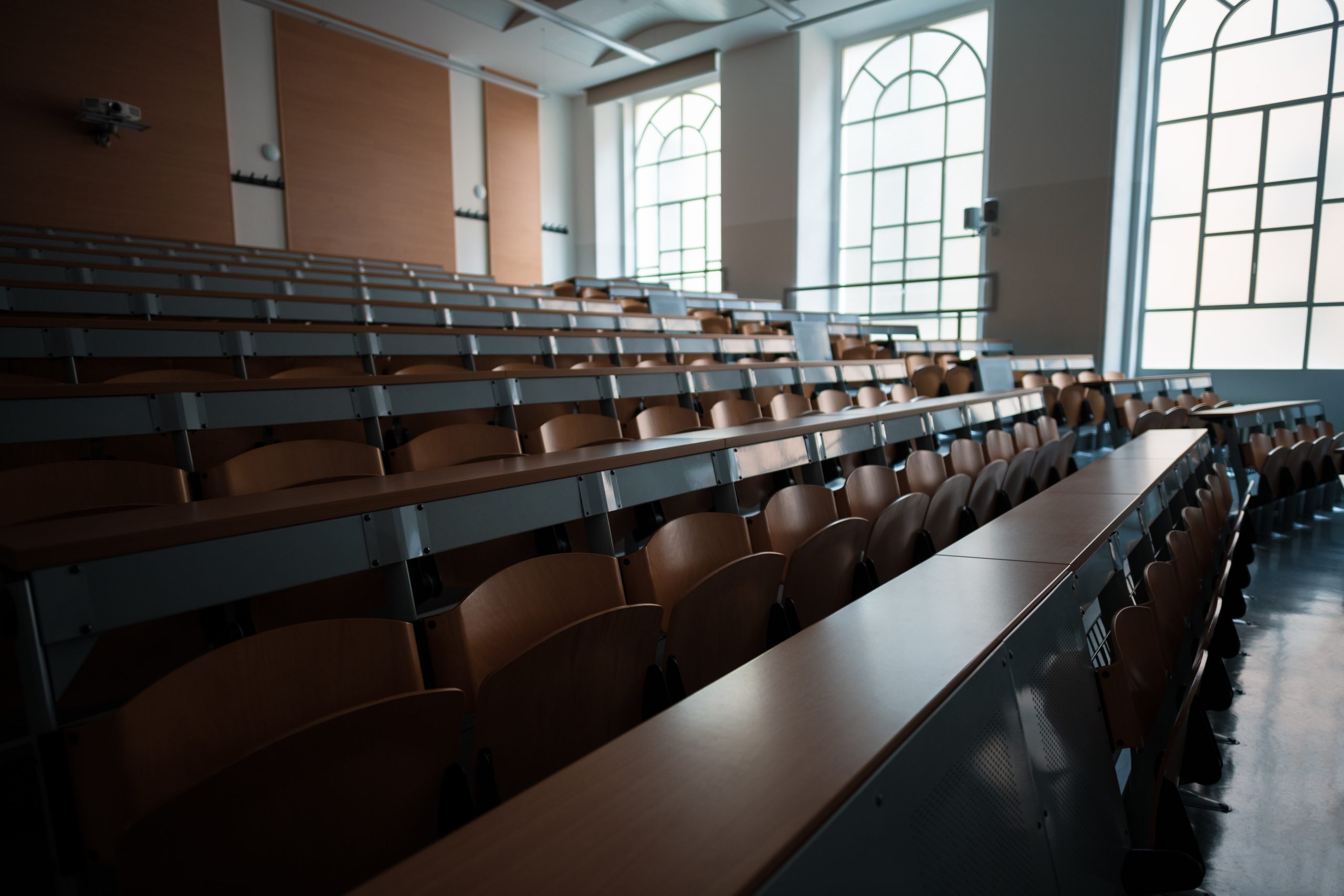 An empty lecture hall with rows of wooden seats and desks, illuminated by natural light coming through large arched windows.