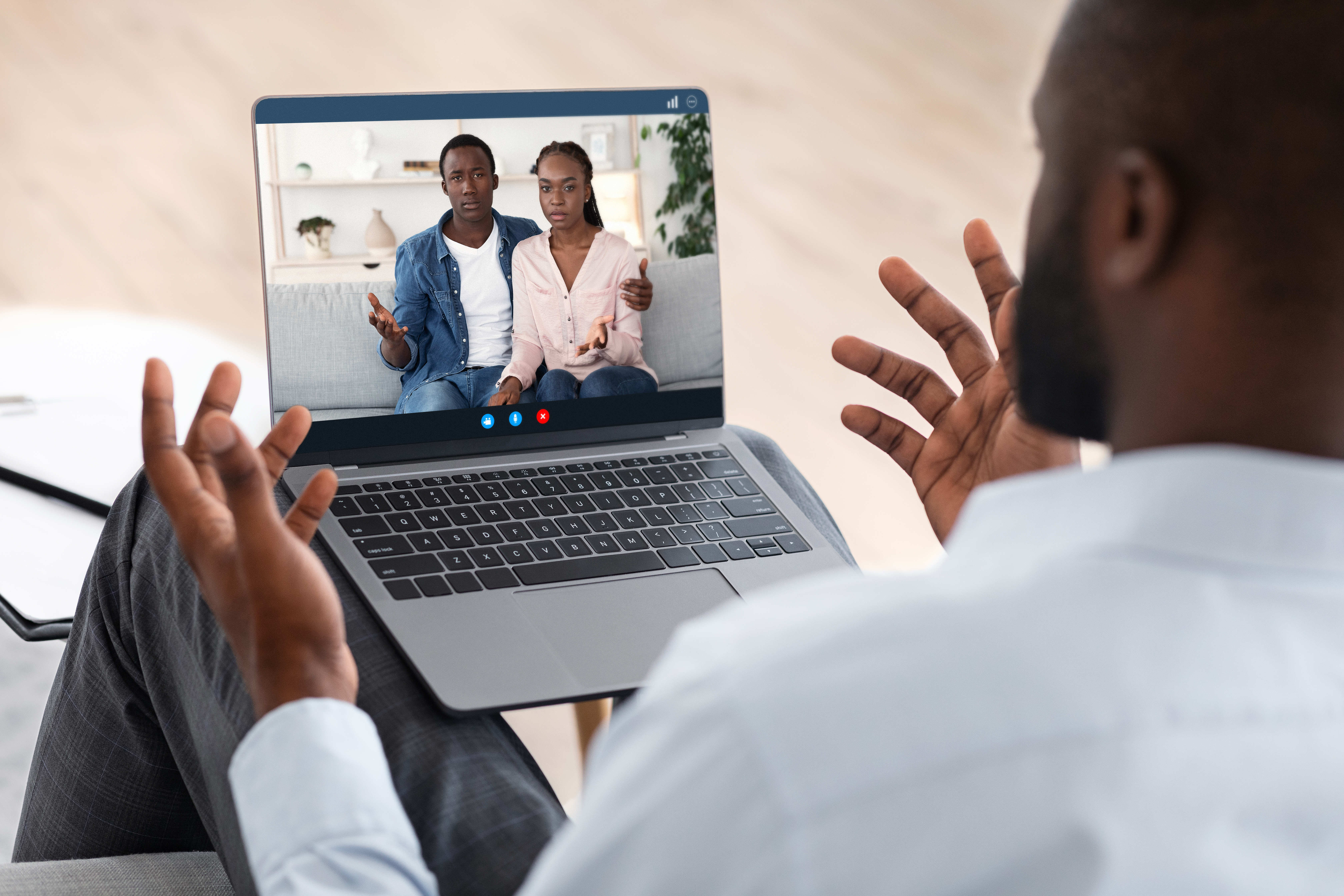 The image shows a person participating in a video call on a laptop. On the screen, there are two individuals sitting on a sofa, engaged in a conversation. The setting appears to be a cozy, well-lit living room with decorative items in the background.