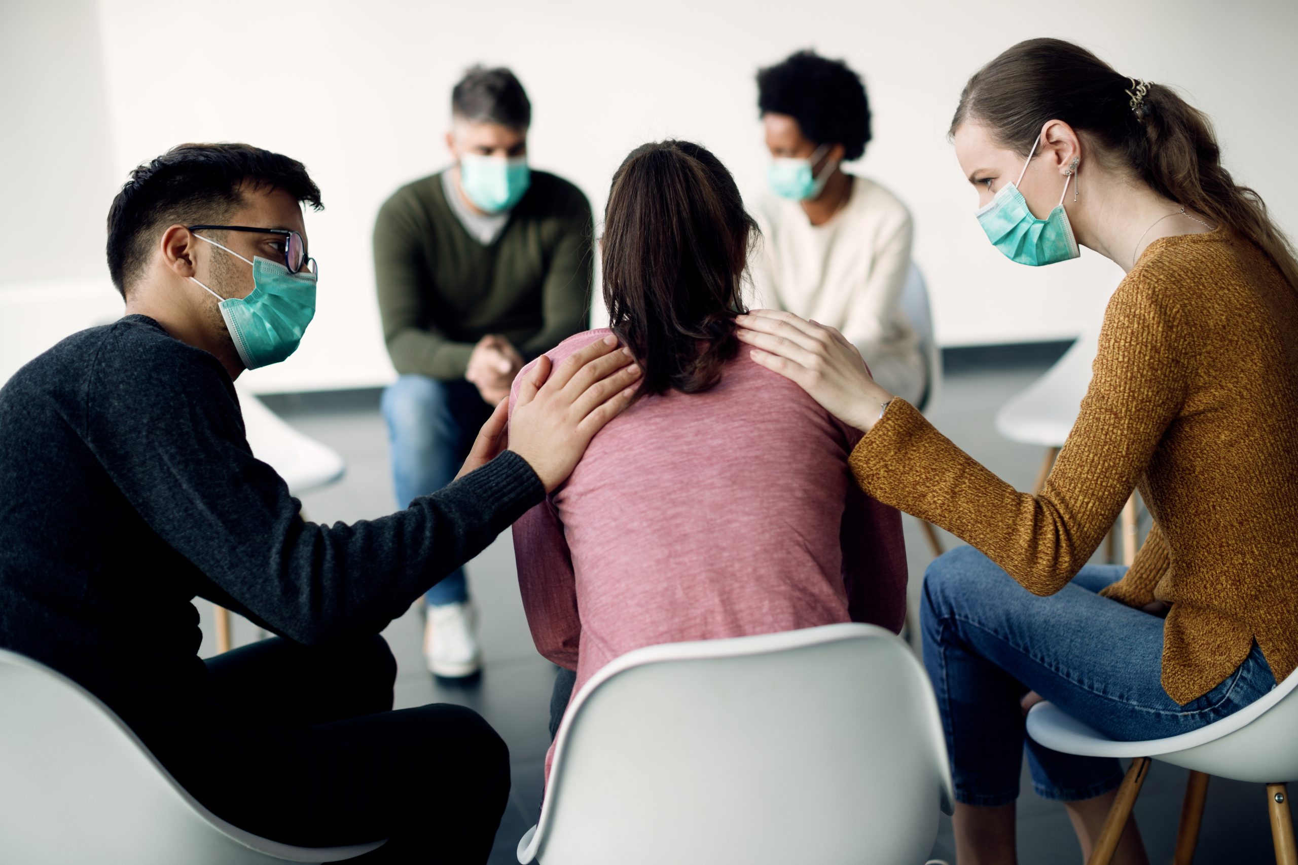 Woman and girl sitting with hands on the girl's shoulders, all wearing masks, in a support group setting.