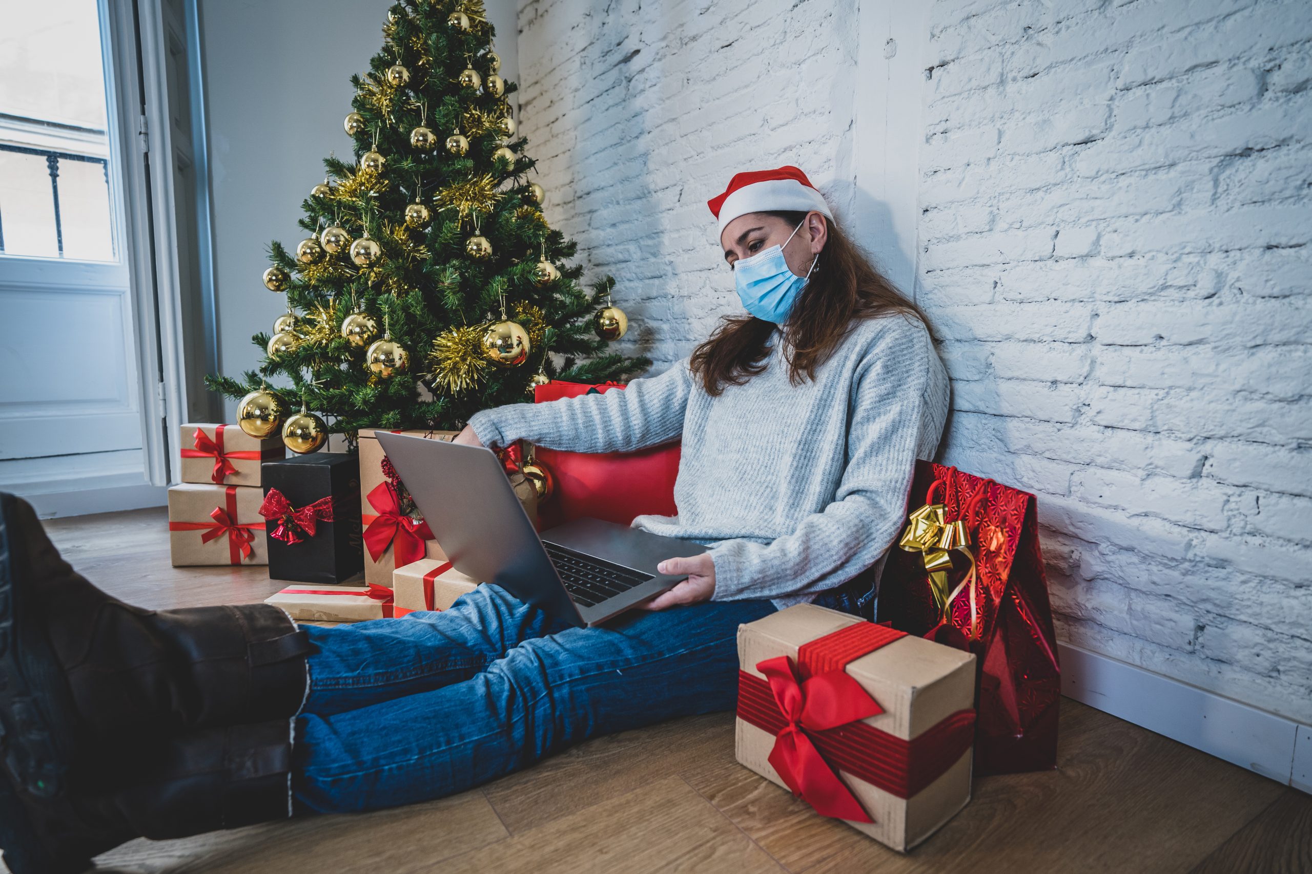 A woman wearing a face mask and a Santa hat sitting on the floor next to a Christmas tree, surrounded by wrapped presents, while using a laptop.