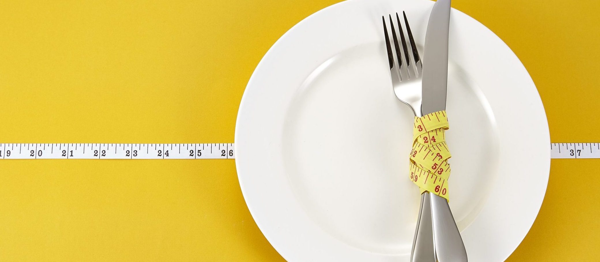 A white plate with a fork and knife crossed on top, wrapped together with a yellow measuring tape, placed on a yellow background with a measuring tape running horizontally across the image.