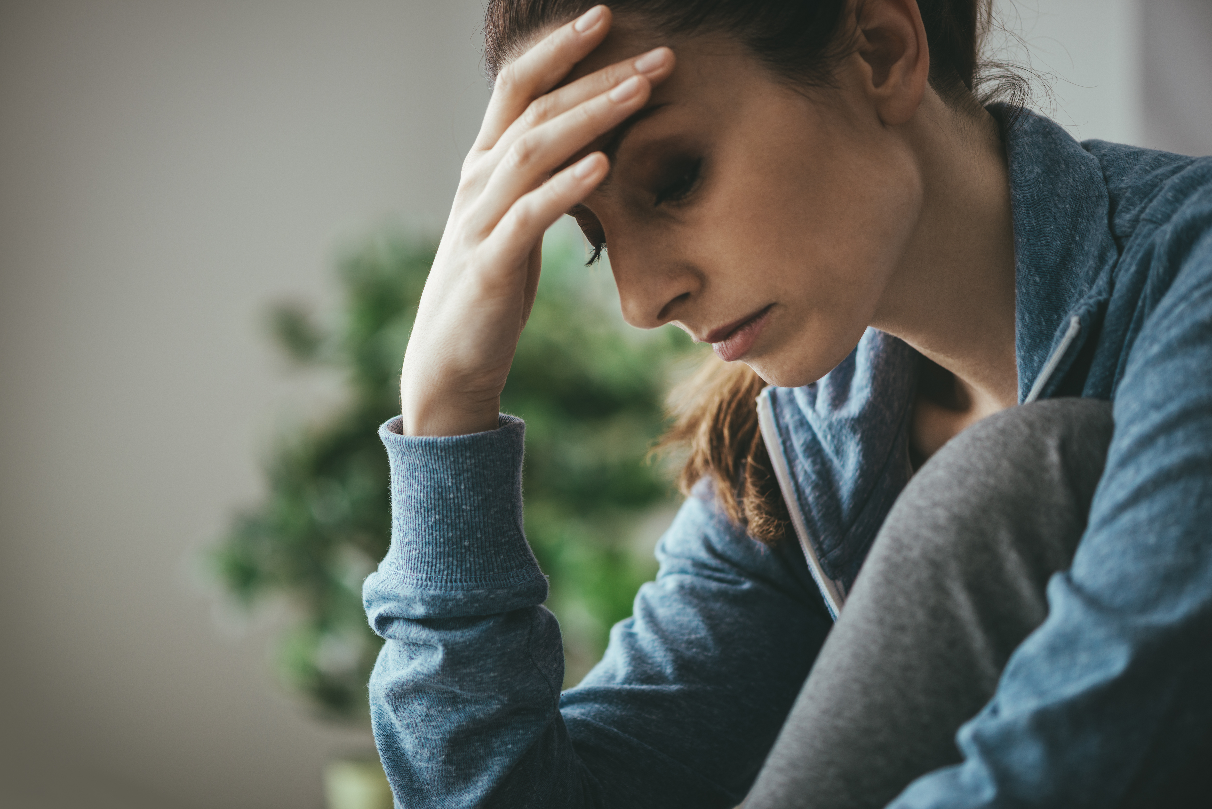 Woman sitting with her head resting on her hand, looking down in sadness or emotional overwhelm, wearing a blue sweater.
