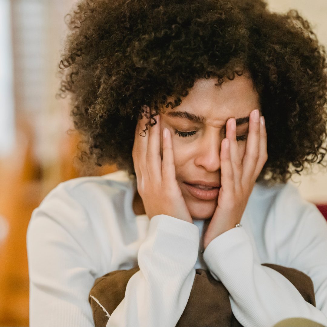 A lady with curly hair stressed out with her eyes closed and her hands over her face.