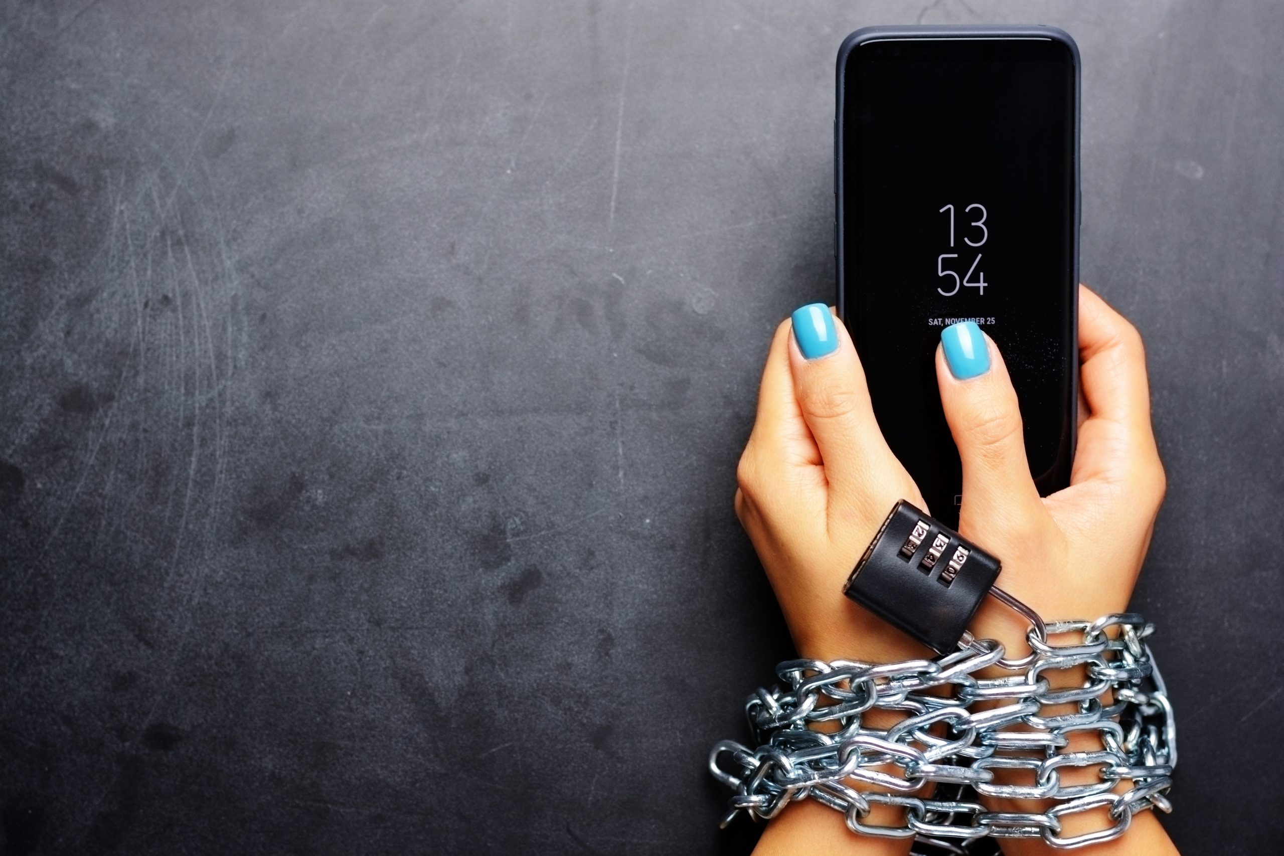 A hand with blue nails is holding a smartphone displaying the time 13:54 and the date Saturday, November 25. The wrist is chained with a metal chain and has a black combination lock.